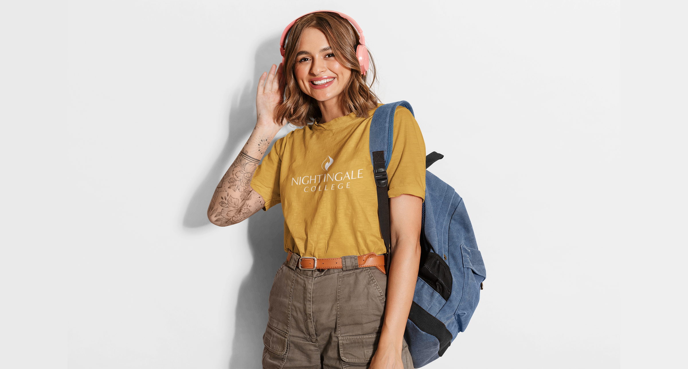 Girl in gold shirt with Nightingale College logo on front wearing a blue backpack with pink headphones in front of cream background